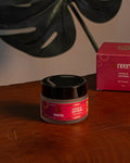 A small cosmetic jar with a black lid and pink label sits on a wooden table next to its pink box. A large dark green monstera leaf decor element is mounted on the wall behind them against a neutral beige background.