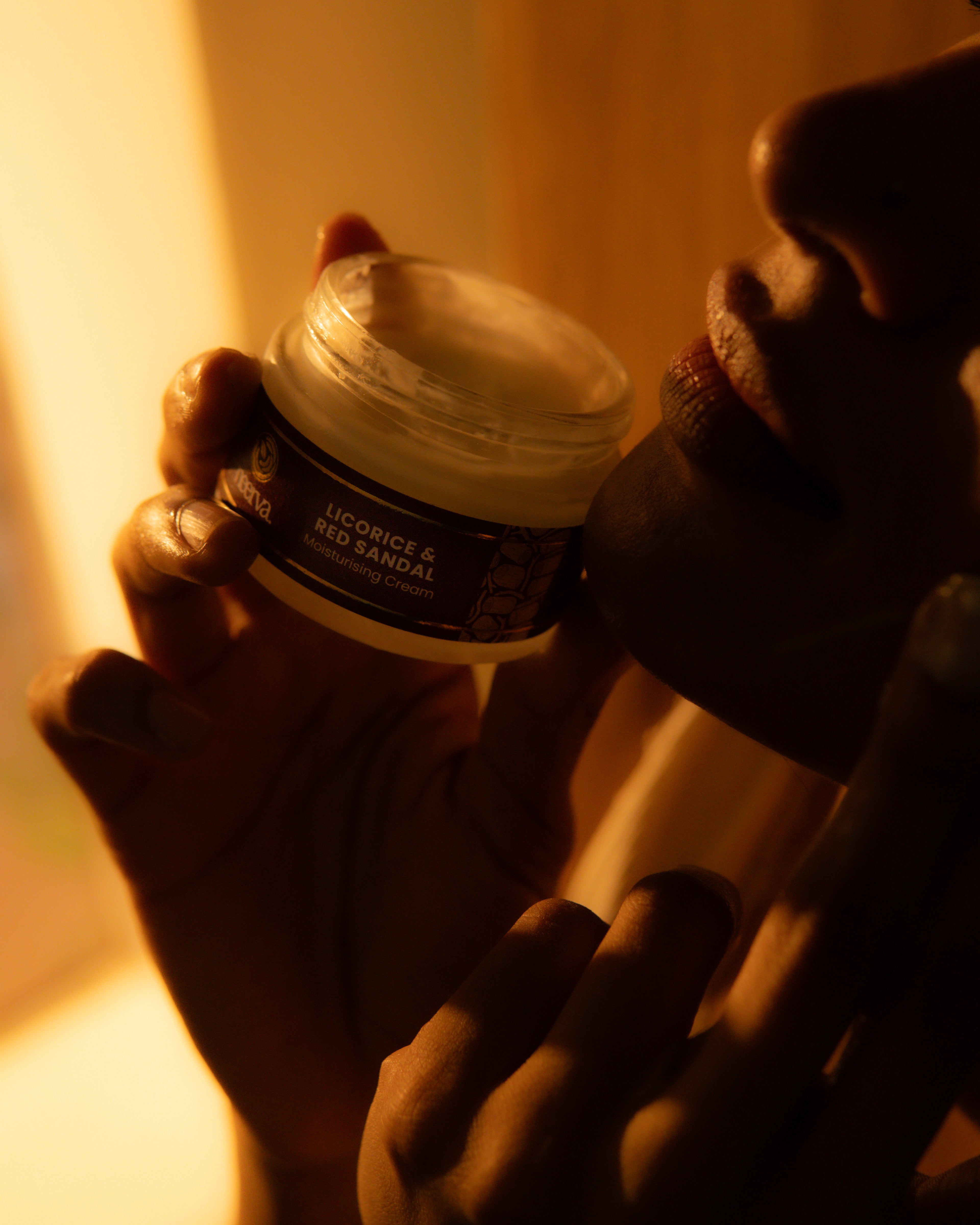 A person holds an open jar of Neerva moisturising cream labeled 'LICORICE & RED SANDAL' against warm, dimly lit skin.