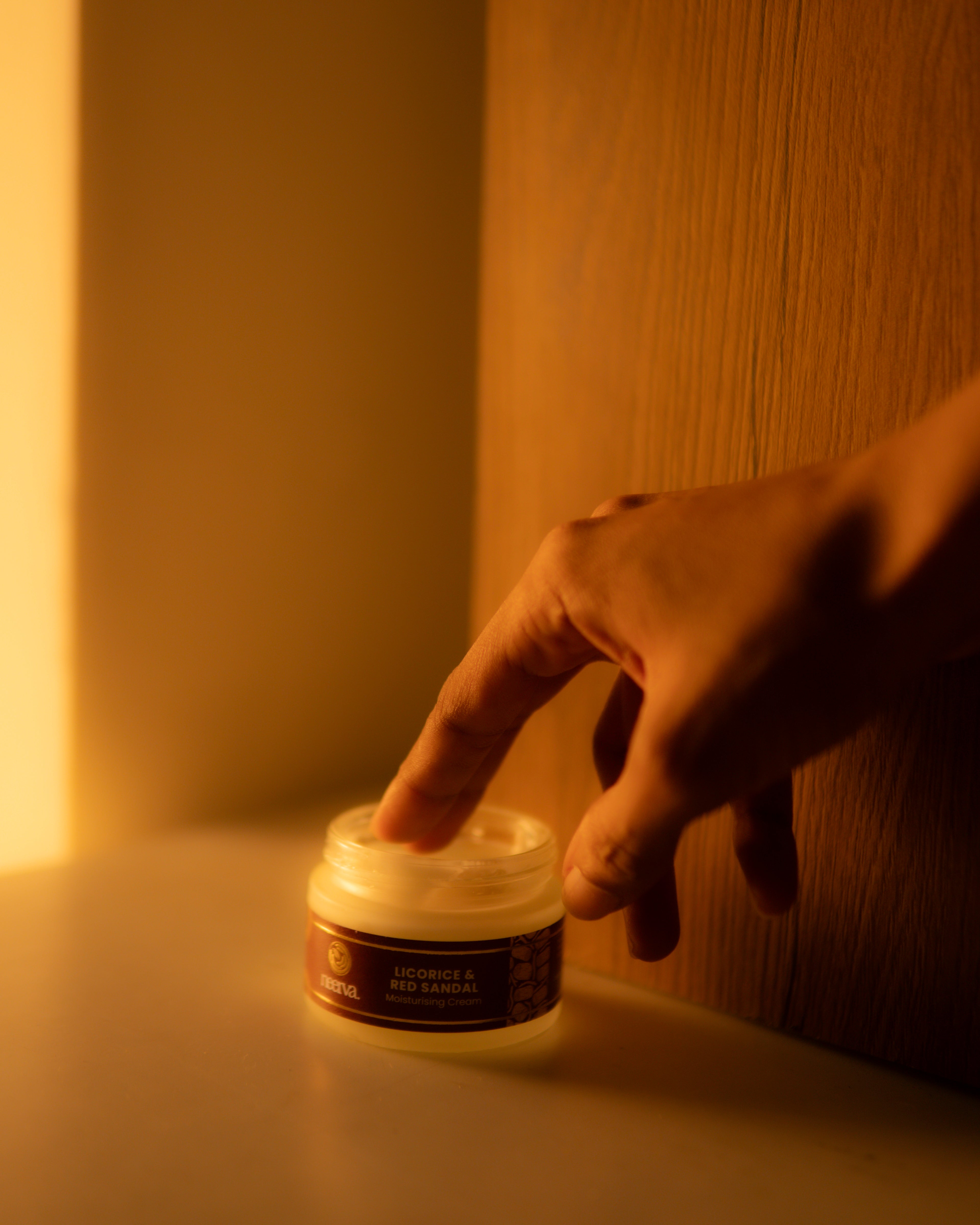 A hand reaching toward a jar of Neerva Licorice and Red Sandal Moisturising Cream on a tabletop in warm, dim yellow lighting.