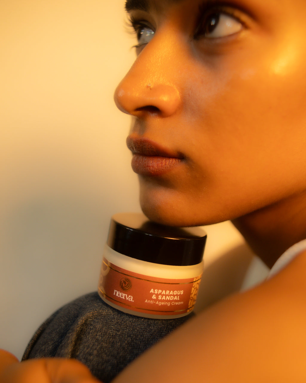 A close-up lifestyle shot of a model resting their head on a gray blanket, with a jar of Reevyl Asparagus & Sandal anti-aging cream on their shoulder.