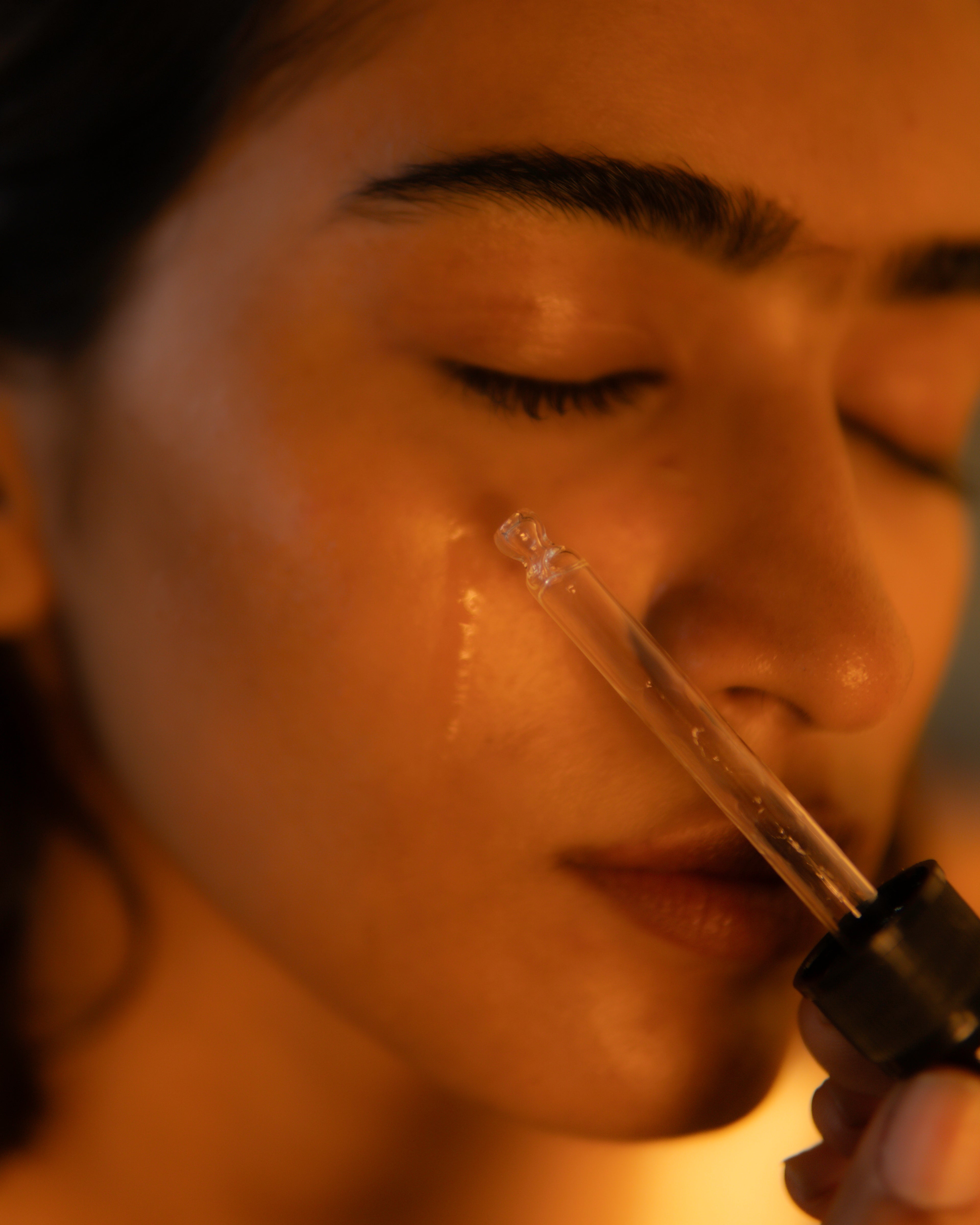 Close-up of a woman's face with a clear dropper applying a glossy liquid to her cheek in warm golden lighting.