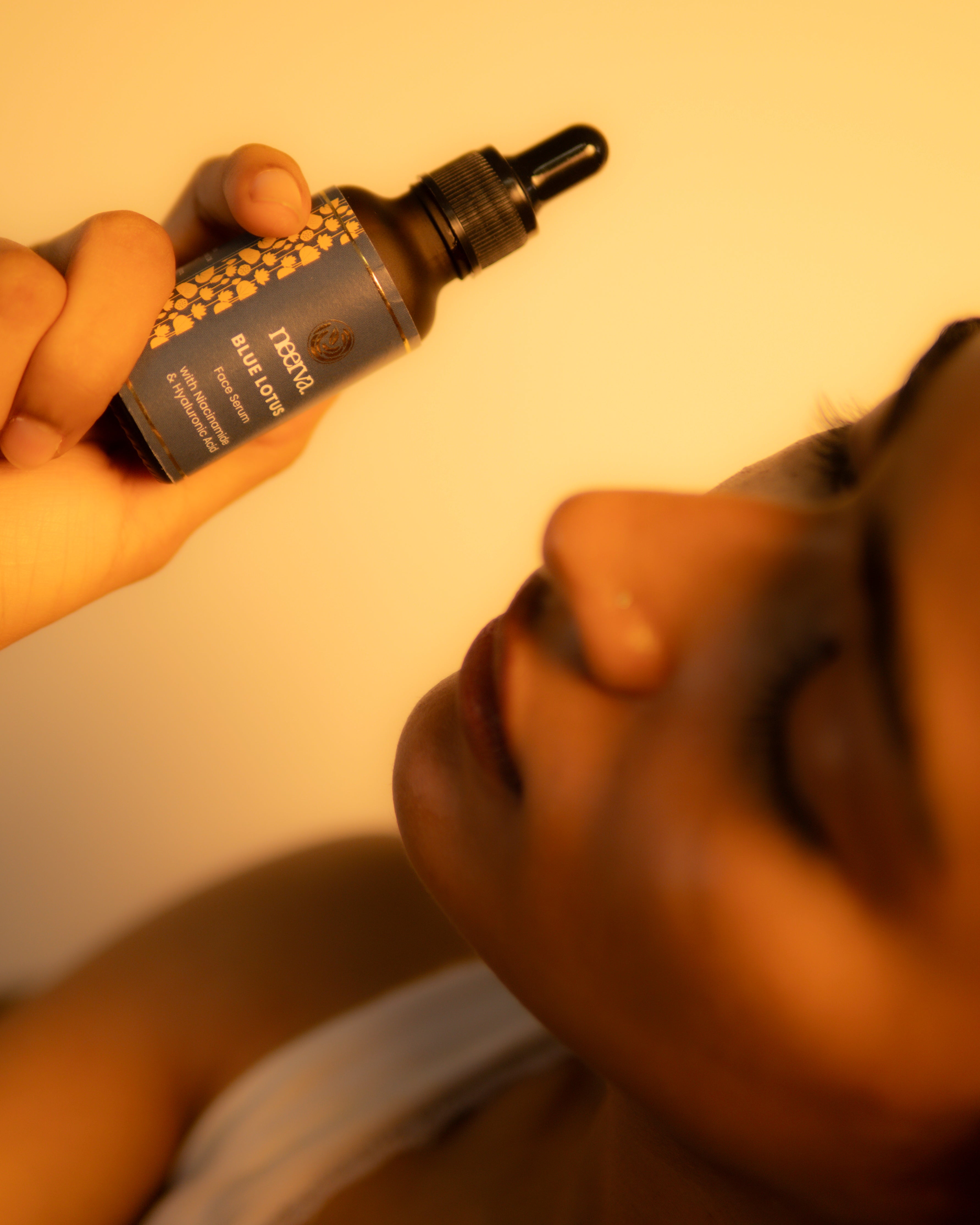 A hand holds a dark amber dropper bottle labeled Neerva Blue Lotus near a person's face against a warm yellow background.