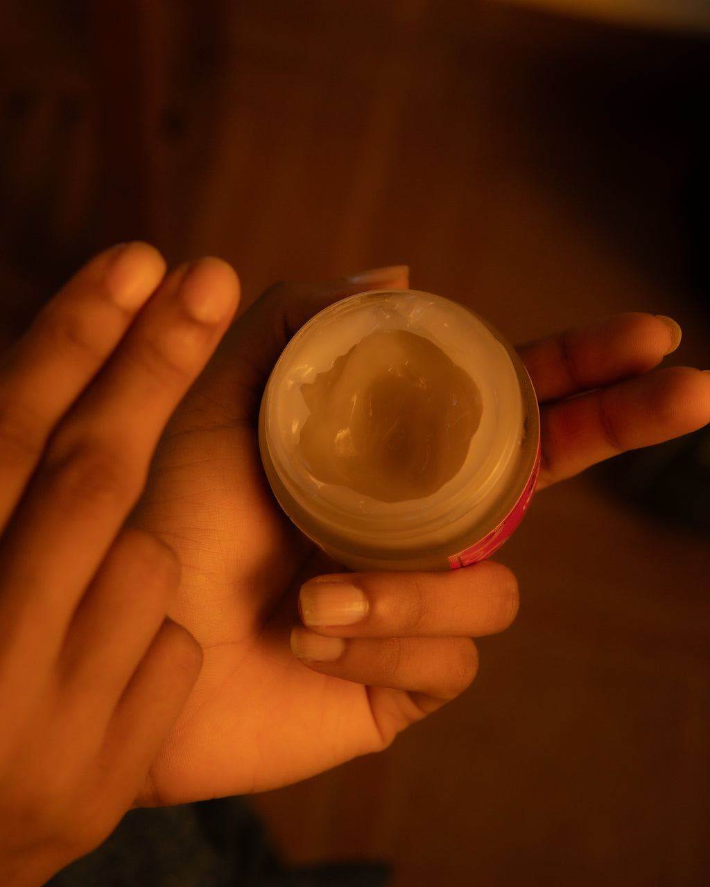 A hand holding an open jar of cream or balm, showing the pale product inside, in warm, dim lighting against a dark brown background.