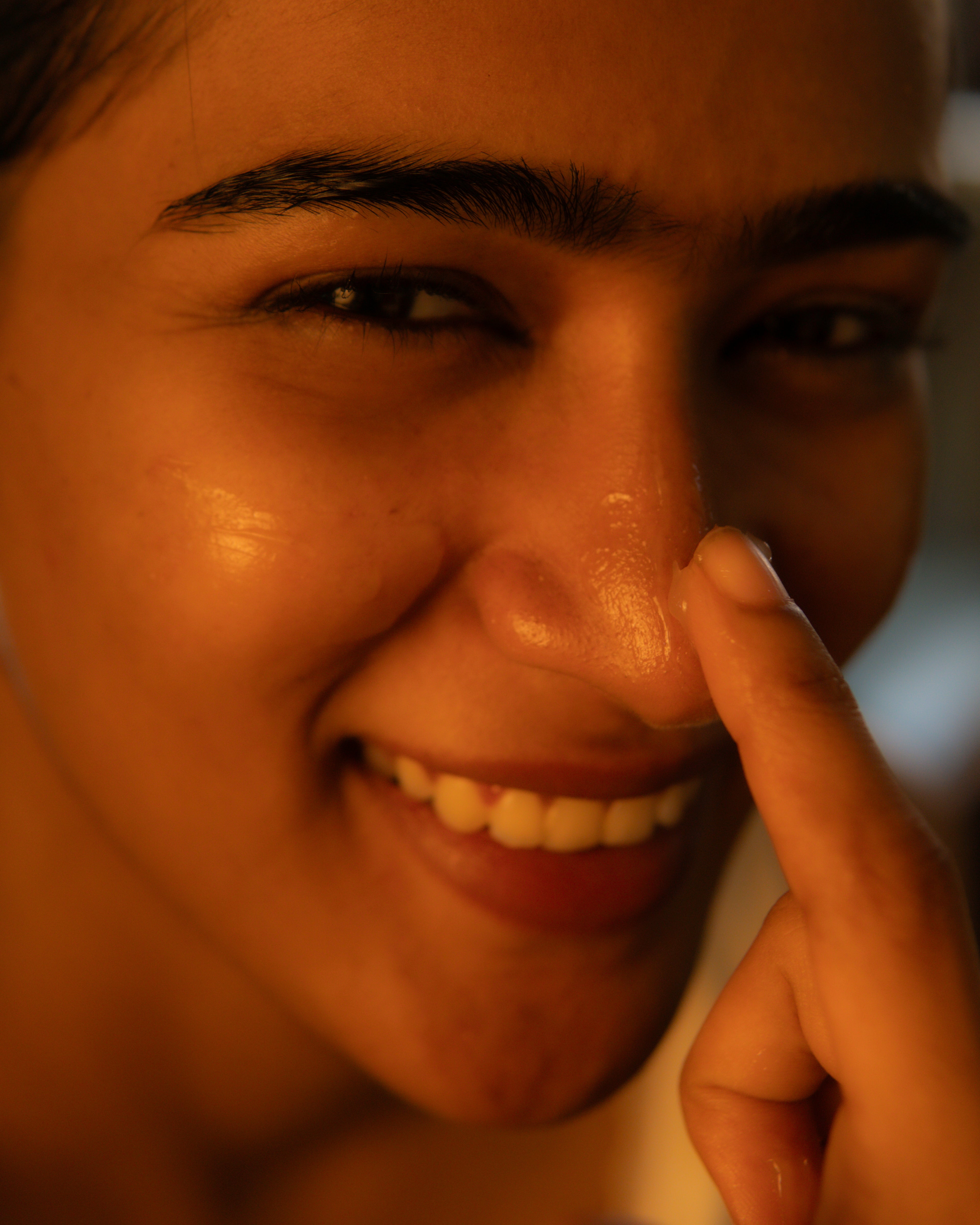 A close-up shot of a smiling woman touching her nose with her finger, showing a glossy, hydrated skin texture under warm, golden lighting.