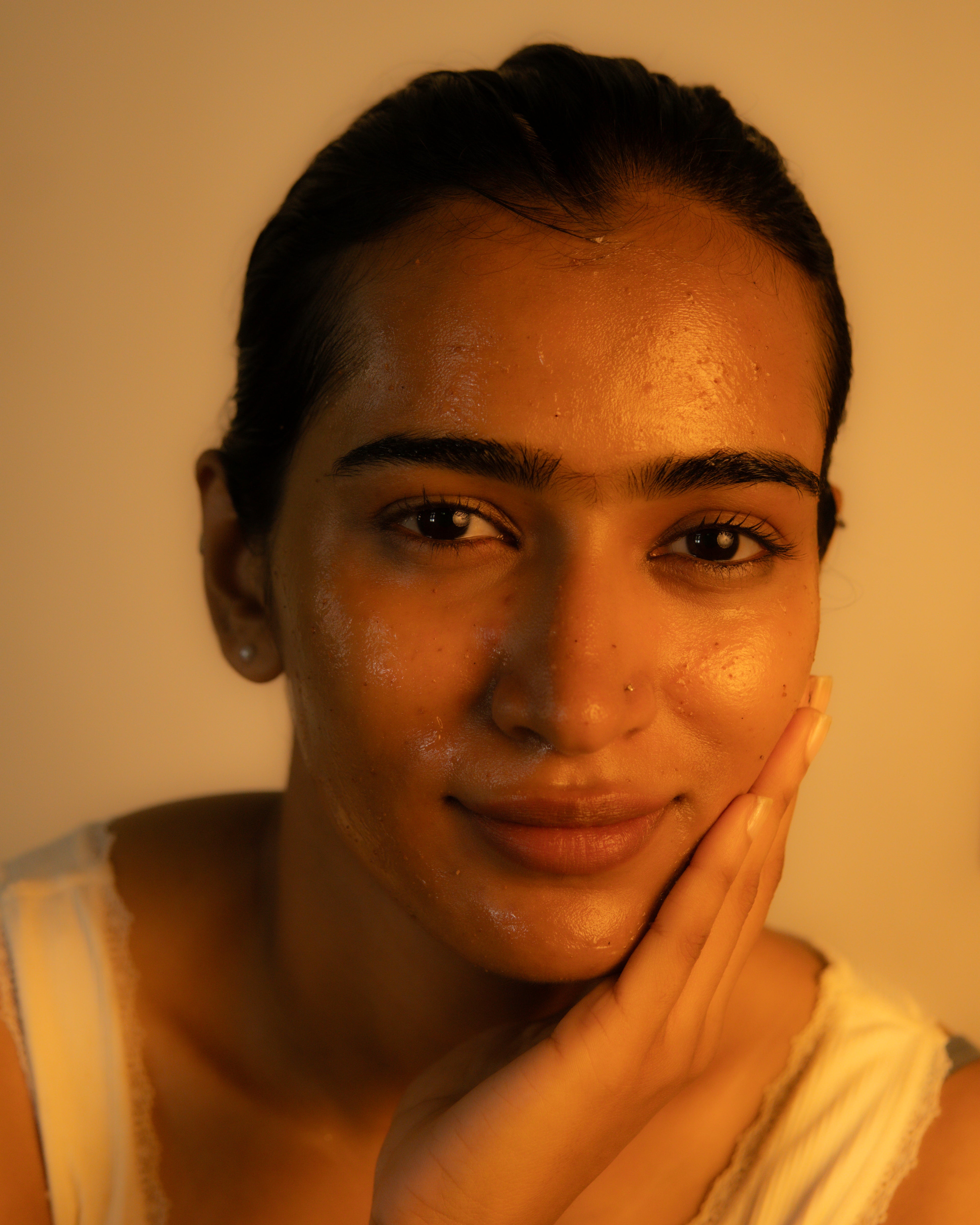 A close-up portrait of a woman with a glossy, hydrated complexion resting her head on her hand, wearing a white tank top against a warm, yellow-toned background.