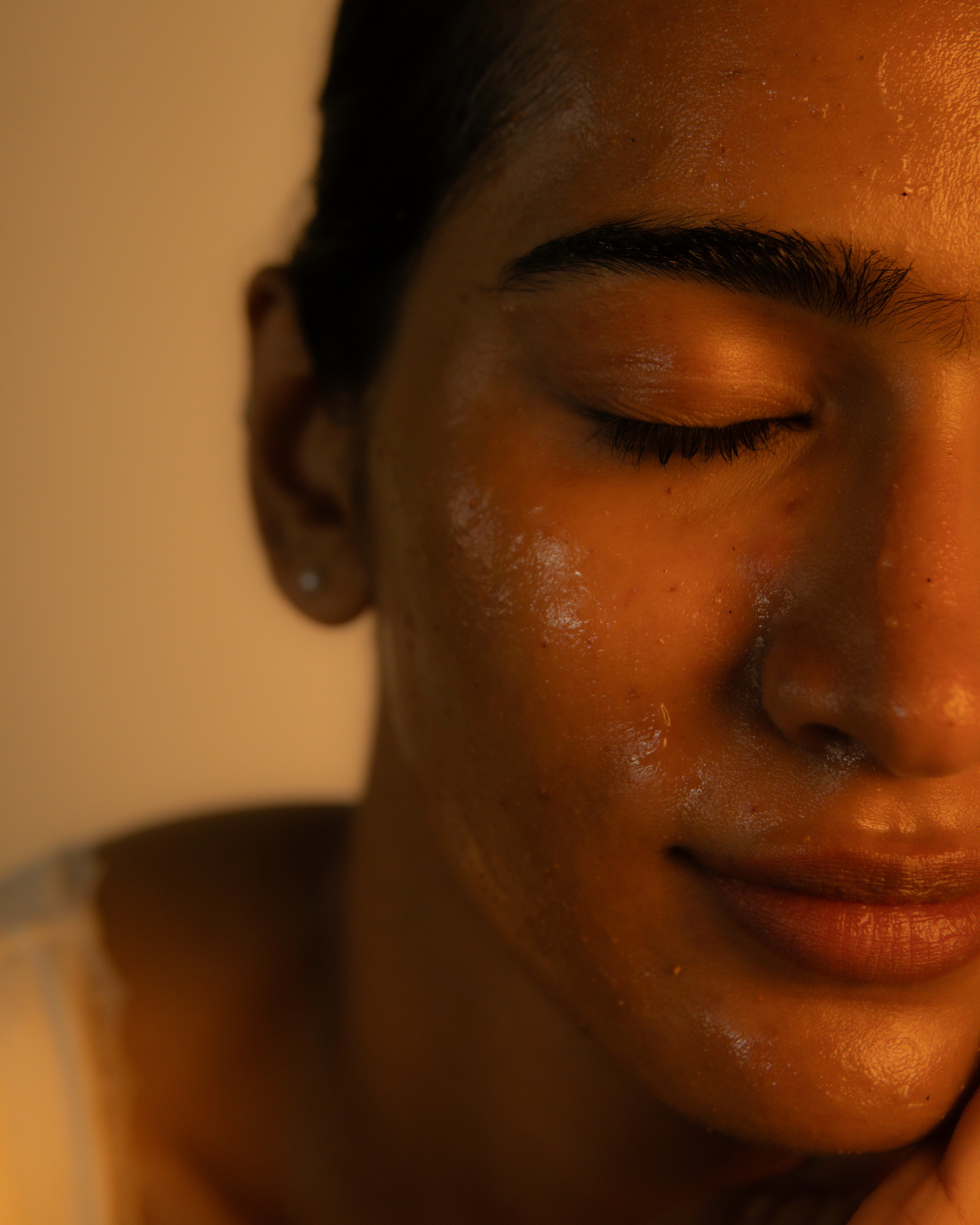 Close-up of a woman's face with closed eyes, wearing a clear gel or oil across her forehead, cheeks, and nose, in warm dim lighting against a beige background.