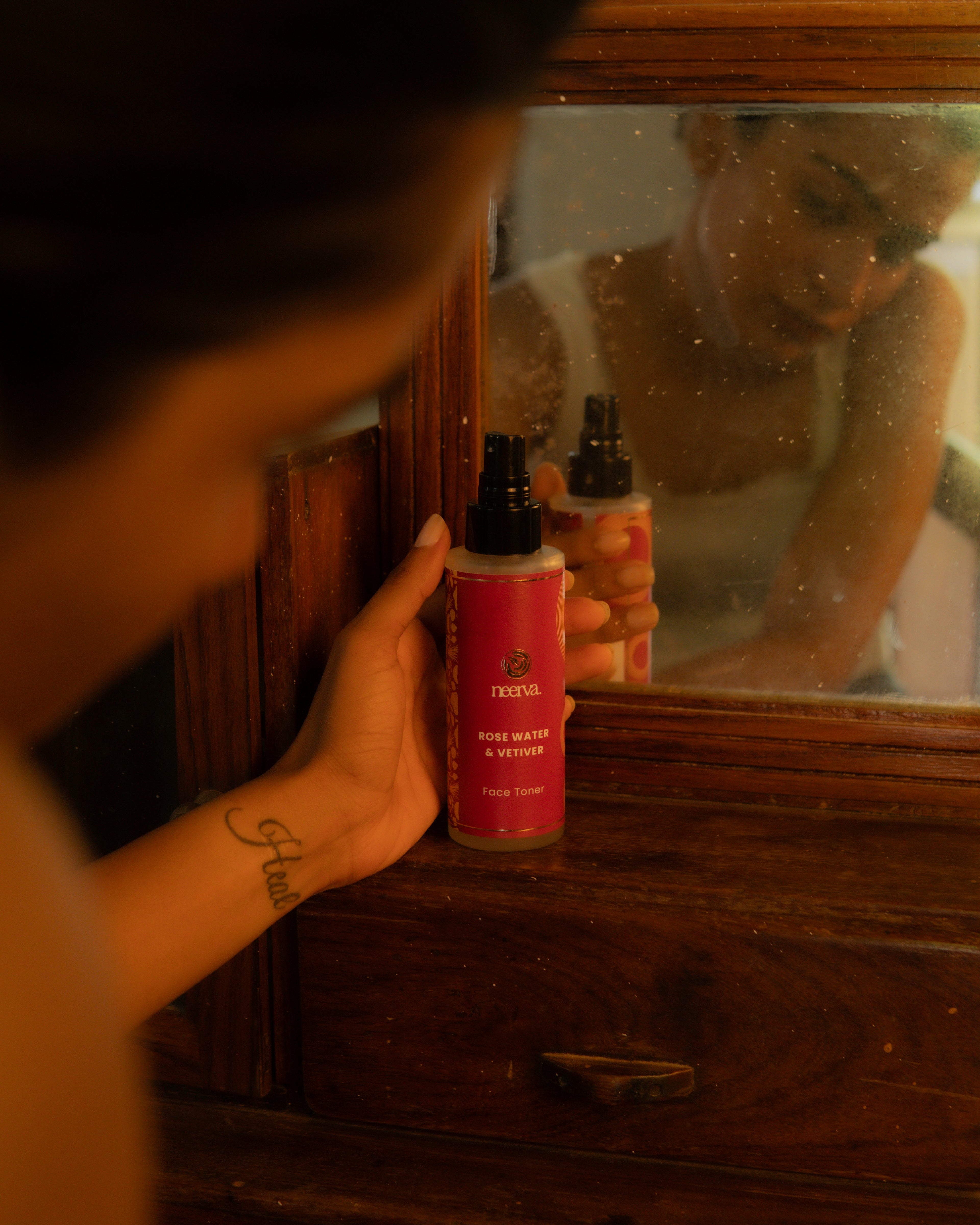 A person holds a bottle of Neerva Rose Water & Toner next to a mirror, showing their reflection while applying or spraying the product. The scene is shot in a dim, warm, rustic indoor setting with a dark wooden frame.