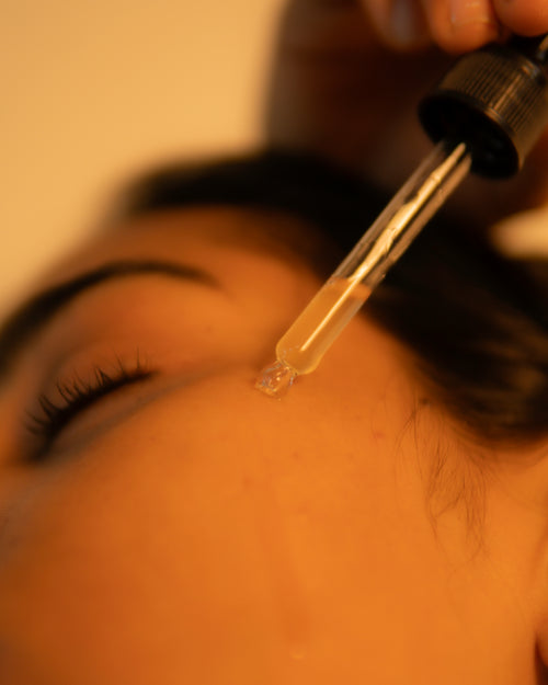 A close-up of a clear glass dropper holding a small drop of transparent liquid above a person's closed eye, with warm yellow lighting.