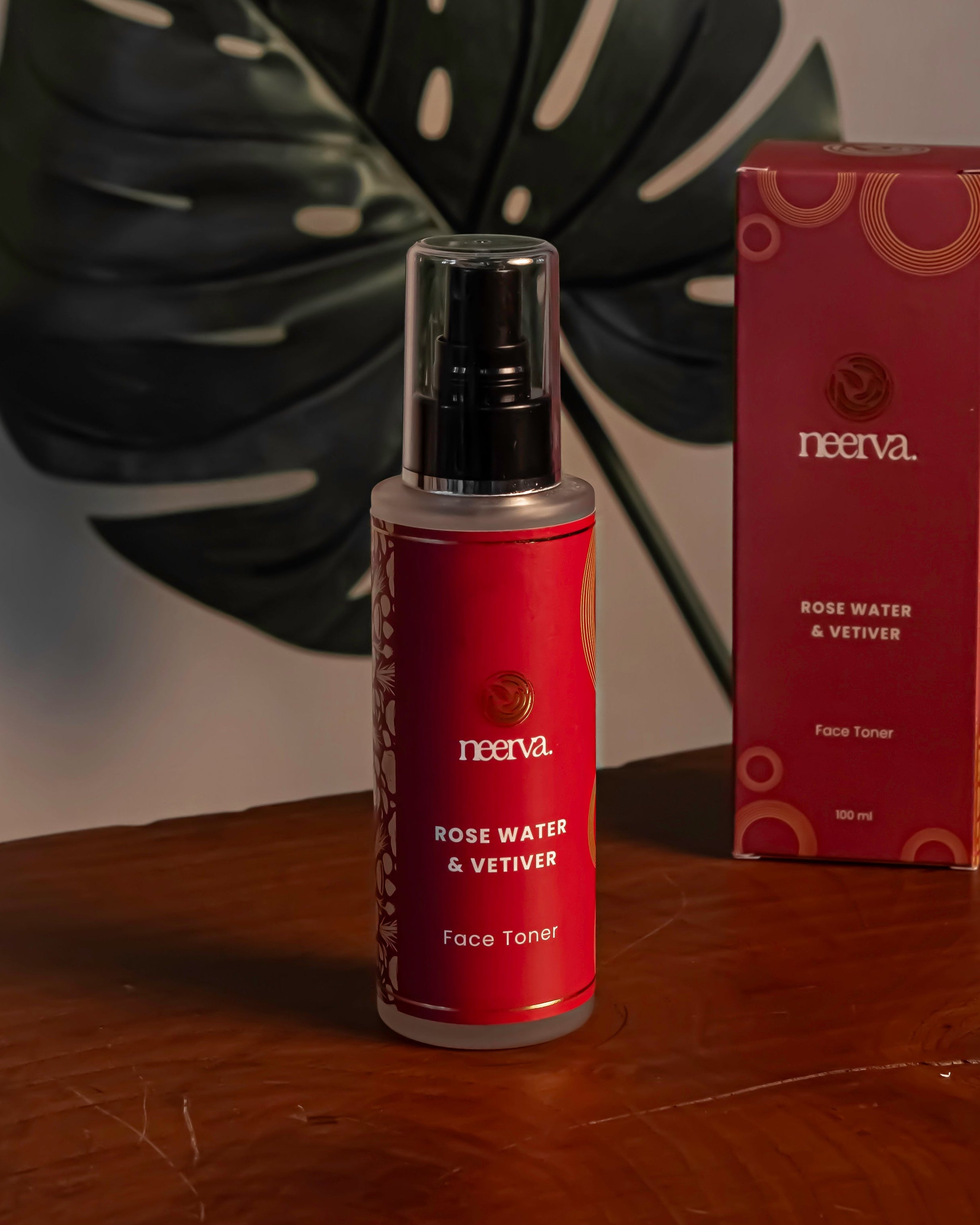 A bottle of Neeva Rose Water & Vetiver Face Toner with a black spray cap stands next to its red packaging box on a polished wooden table, with a large green monstera leaf in the background.
